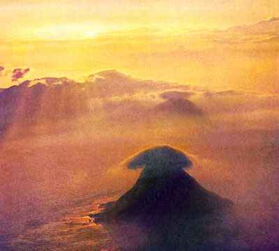 A golden sunset over a cloudy sea; steep conical islands, each with a dark lenticular cloud for a cap.